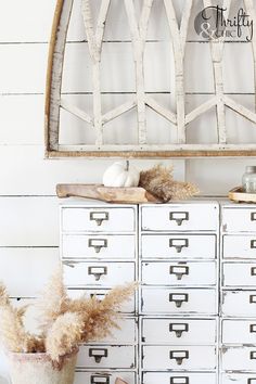 a white dresser with drawers and plants on it in front of a wooden wall hanging