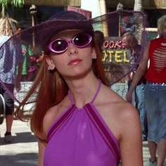 a woman in a purple dress and hat on the street with people walking behind her