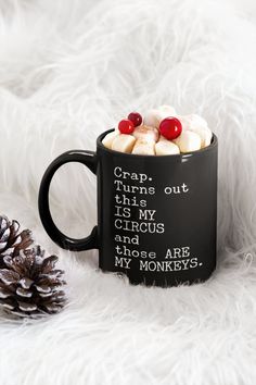 a black coffee mug with marshmallows and candy on it sitting next to a pine cone