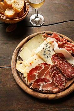 an assortment of cheeses, meats and bread on a wooden plate next to a glass of wine