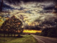 the sun is setting on an empty road with trees in the foreground and clouds in the background