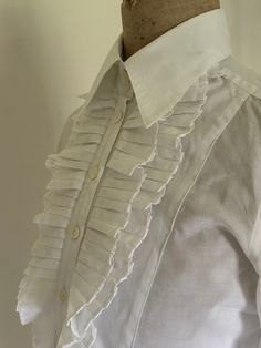 A fabulous vintage white cotton blouse with a pleated front frill / bib , shaped seam details and lovely mother of pearl buttons down the front and on each cuff.  It has a deep collar band and a pointed collar. There is also a lovely detail on the front button placket where a small tab is incorporated into the placket and fastens with one of the buttons. Length : 21 1/2" Chest : 34"  Hem circumference: 34" Underarm : 18" Cuff depth : 3 3/4" Cuff circumference: 8" Collar band : 2" Collar depth at Classic Office Shirt With Ruffled Collar, Classic Shirt With Ruffled Collar For Office, Classic Ruffled Shirt For Office, White Ruffled Shirt For Formal Occasions, Formal White Ruffled Shirt, Classic Cotton Blouse With Ruffles, Formal Ruffled Collared Shirt, Classic Shirt With Ruffled Collar For Work, Classic Shirt With Ruffled Collar For Formal Occasions