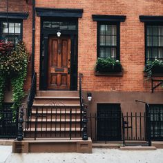 a brick building with black iron railings and wooden front door, surrounded by green plants