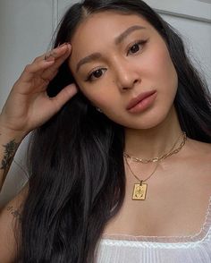 a woman with long black hair wearing a white top and gold necklace holding her hand up to her ear