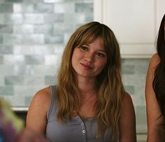 two young women sitting next to each other in a kitchen, one is looking at the camera