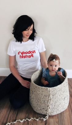 a woman sitting on the floor next to a baby in a basket with a t - shirt that says nobody's perickt