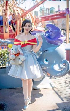 a woman in a dress standing next to an elephant at a amusement park with her stuffed animals