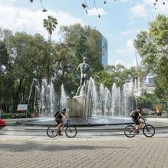 two bicyclists ride past a fountain in a park