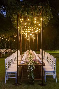 an outdoor dinner table with white chairs and lights hanging from the ceiling over it, surrounded by greenery