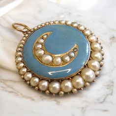 Late Georgian / Early Victorian Enamel and Pearl Moon Locket in 14K Gold This exquisite antique locket, beautifully crafted in 14K gold, features an enchanting crescent moon motif on its front. The moon, inlaid with lustrous pearls, sits against a backdrop of soft, sky-blue enamel, giving the piece an ethereal charm. The outer edge of the locket is framed with additional pearls, enhancing its timeless elegance. The back of the locket opens to reveal a secure glass closure, perfect for holding cherished mementos, such as photographs or small keepsakes, allowing you to keep your loved ones or memories close. With its celestial theme and delicate craftsmanship, this locket is a stunning piece of history and a unique accessory for any collection. Please note that antique and vintage jewelry ma Antique Evening Locket Jewelry, Victorian Ceremonial Locket Jewelry, Antique Enamel Locket Jewelry, Victorian Enamel Locket Jewelry, Victorian Brass Locket Necklace Collectible, Antique Locket, Vintage Fans, Locket Necklace, Accessories Unique