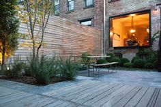 an outdoor patio with tables and benches next to a brick building at night, surrounded by greenery