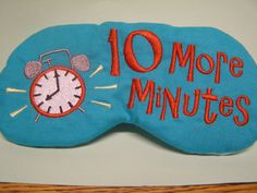 "Alarm Clock with the words \"10 more minutes\" embroidered in red on teal/aqua cotton fabric. Alarm clock is red, black and gray with yellow accents.  Black non-adjustable strap, black cotton flannel back. Packaged in a cello bag  Order as shown and described OR CREATE YOUR OWN MASK - Change design colors and fabric colors (over 100 color choices each!) - Select blue star or black flannel back  - Elastic strap keeps it secure (adjustable strap also available) - Mask is approx. 6.5\" across, approx. 3\" tall (Mask fits 20-22\" head) - Strap is approx. 13.5\"  - Order with or without lavender (USA only) - Light blocking fabric panel and 3 layers of batting - Pillow softness - Perfect for adults and kids (age 5+ recommended) and adults - Machine wash/dry (Only for masks WITHOUT lavender) - S Clock Eye, Cute Sleep Mask, Cute Sleep, Eye Mask Sleep, Guys Night, Tissue Case, Black Flannel, Shopping Spree, Sleep Mask