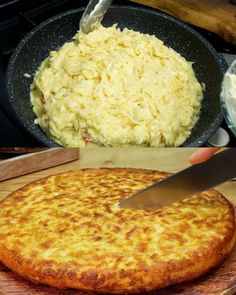 someone is cutting into a cheese pizza on a wooden board next to a skillet full of mashed potatoes