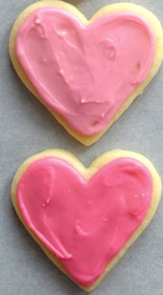 two heart shaped cookies with pink icing on a gray surface next to each other