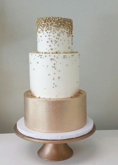 a three tiered cake with gold sprinkles sits on a white table