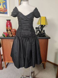 This is a beautiful show stopping well made 80s cocktail dress by an English label that I think says Carmonia Collection in a size 12uk The fabric is a swishy black tafetta like fabric ( actually polyester) and  fully lined. The dress has a wide neckline that sits on the edge of the shoulders with little pleated cap sleeves and a fitted rouched  bodice and a dropped waist. The skirt is gathered with a tuile underskirt between the outer and the acetate lining. No prickles.  The condition is very Vintage Formal Dress With Ruched Bodice, Vintage Evening Dress With Ruched Bodice, Fitted Party Dress With Gathered Skirt, Fitted Dress With Gathered Skirt For Party, Black Party Dress With Gathered Skirt, Fitted Evening Dress With Pleated Waist, Vintage Pleated Midi Dress For Evening, Fitted Evening Dress With Pleated Waist For Cocktails, Black Midi Dress With Gathered Waist For Evening