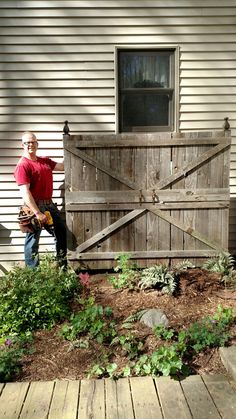 a man standing in front of a wooden door next to a garden filled with plants