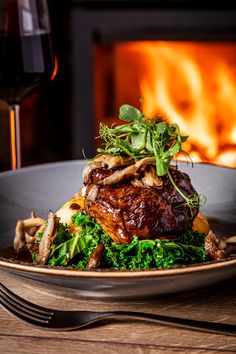 a plate with meat and vegetables on it next to a glass of wine in front of an open fire