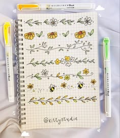 an open notebook with flowers and bees on it next to two markers, one green pen and the other yellow marker