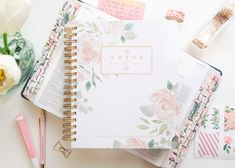 an open planner book sitting on top of a white table next to flowers and pens