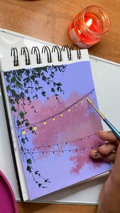 a person is holding a pen and drawing on a notebook with string lights in the background