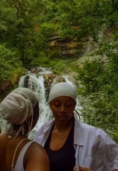 two women standing in front of a waterfall with trees and bushes behind them, one woman has her hand on the shoulder of another woman's back