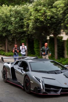 a silver sports car driving down a street with people standing on the side of it