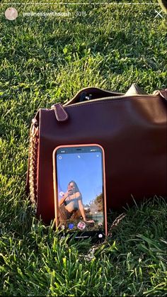 a cell phone laying on the ground next to a brown handbag and purse with a woman's reflection in it