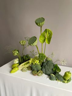 an arrangement of green vegetables on a white table cloth with leaves and flowers in the center