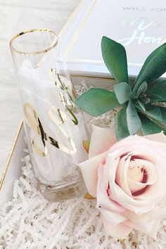 a white rose sitting next to a glass vase with greenery on it and a personalized note