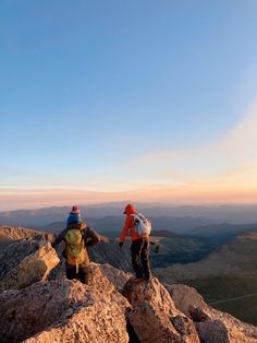 two people standing on top of a large rock in the middle of a mountain range