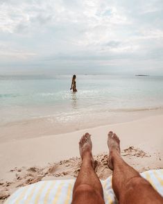 a man is sitting on the beach with his feet in the water and there is a person standing in the ocean behind him