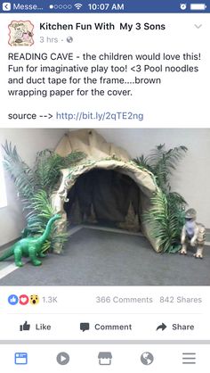 two toy dinosaurs are sitting in front of a fake cave with plants and rocks on the floor