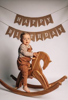 a baby sitting on top of a wooden rocking horse with happy birthday banner in the background