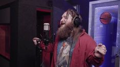 a bearded man with headphones on singing into a microphone in front of a recording booth