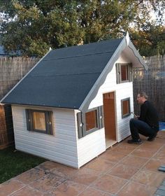 a man sitting on the ground in front of a small white house with a black roof