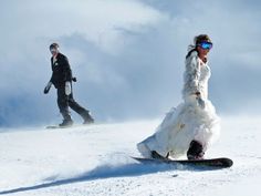 two people are snowboarding down a snowy hill in the day time and one person is wearing goggles