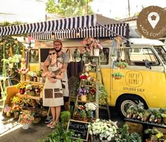 a man and woman standing in front of a yellow van with flowers on display at an outdoor market