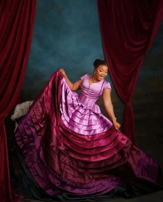 a woman in a pink and purple dress is posing for a photo with red curtains behind her