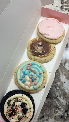 four decorated cookies in a white box on a marble counter top with pink and blue frosting