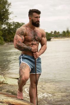 a shirtless man standing on top of a surfboard next to the ocean holding a stick