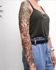 a woman with a tattoo on her arm standing in front of a white wall wearing jeans and a green tank top