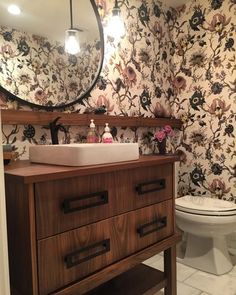 a bathroom with floral wallpaper and wooden vanity, mirror above the sink, and toilet