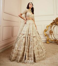 a woman standing in front of a mirror wearing a white and gold lehenga