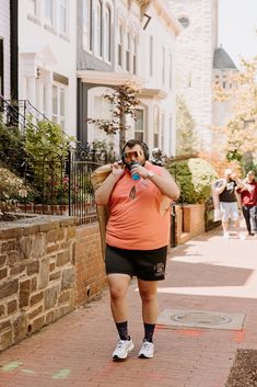 a man in an orange shirt and black shorts is walking down the street with his cell phone to his ear