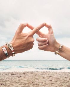 two people making a heart shape with their hands on the beach near the ocean and sky