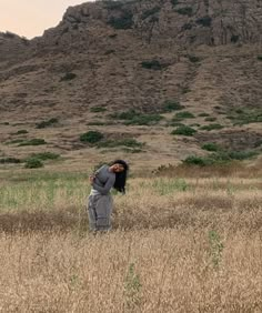 a woman standing in the middle of a field next to a large rock formation on top of a hill