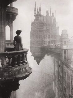 a woman standing on top of a balcony next to a tall building with spires