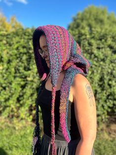 a woman wearing a multicolored crocheted scarf and hat in front of bushes