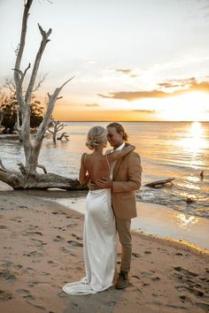 a bride and groom standing on the beach at sunset